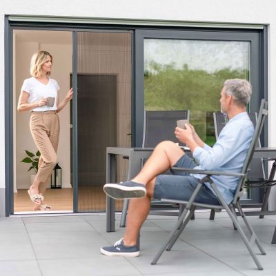 Eine Frau öffnet ein Fliegengitterplissee an der Terrassentür. Ein Mann sitzt im Stuhl und beobachtet sie.